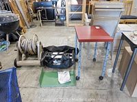 Photo shows Cuisinart portable gas grill, garden hose reel, metal table, and a small black stand in a warehouse setting.