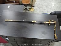 Full-length view of parade sword sheathed in black painted scabbard with brass fittings and gold tassel on black table.