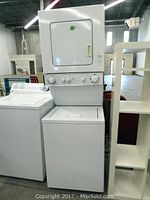 Front view of stackable washer and dryer unit inside a warehouse or store environment, showing dryer on top and washer below.