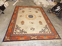 Photo of the full area rug lying flat on concrete floor, showing floral and oriental design with cream center and burnt orange border.