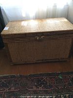 Full view of rectangular wicker bedding box with lid closed, showing natural beige woven wicker texture and metal clasp on the front.