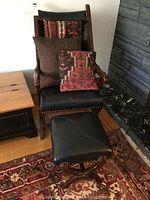 Front view of black pleather armchair with dark wood arms and legs, decorated with patterned cushions (not included), matching stool in front
