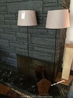 Two black metal floor lamps with taupe shades standing in front of a stone fireplace, black finish, slender stands, large drum shades.