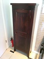 Closed dark wood tall narrow cupboard with single door and round wooden knob, slight scuffs visible.