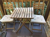 Photo showing wooden slatted patio table with foldable legs, two wooden folding chairs with beige cushions, and a wooden baby gate next to the chairs on a porch deck.
