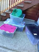 Five clear and colorful plastic storage bins containing assorted Christmas and Halloween decorations and two green metal Christmas tree stands