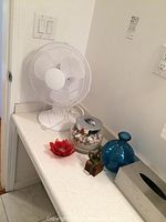 White 19 inch fan, glass jar with sand and shells, red flower petal candle holder, blue vase, metal man sculpture sitting on block on a white countertop.