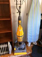 Full view of yellow and gray Dyson DC07 upright vacuum cleaner standing vertically next to furniture. Visible crack on lower gray section. Clear dust bin present.