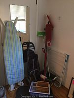 Photo showing ironing board with blue-green-white striped cover, black foldable shopping cart, collapsible drying rack and multiple cleaning tools including mops and broom.