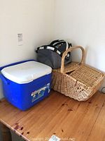 Three items on wooden floor: blue 16 quart Coleman cooler, gray foldable insulated food hamper, and woven picnic basket with a handle.