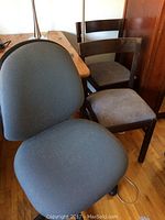 Office swivel chair and two wooden kitchen chairs with grey fabric seats, one wooden chair fully visible with scratches on wood, office chair in front showing ergonomic seat and back.