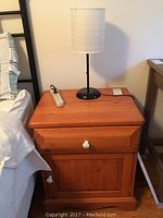 Solid wood night stand viewed from front, showing drawer and cabinet door with white knobs, scratches visible on wood surface, and black base lamp with white paper shade on top.