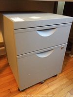 Front angled view of grey metal 2 drawer filing cabinet on castors with lock on lower drawer but no key.