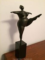 Front angled view of the dark toned composite statue of a dancing lady with one leg raised, arms open, standing on black base