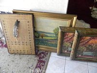 Overview of all three framed pieces leaning against wall; two scenic paintings and one historic printed document with feather