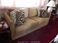 Full view of tan sleeper sofa with four decorative pillows on it, placed in a room with red carpet and pink curtains.