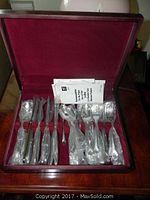 Opened wooden flatware case with maroon velvet lining holding multiple pieces of J.A. Henckels flatware wrapped in plastic.