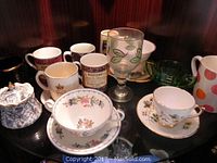 Multiple assorted teacups, mugs, saucers, and glassware arranged on a black surface, showing various colors, designs, and sizes