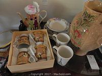 View of the teapot, four decorated mugs in a box, two white mugs, large floral jug, and stack of world map dishes including plates and bowl.