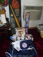 Shadow box containing baseball glove, bat, ball, vintage photos positioned vertically behind a collection of Yankee cups, mugs, and a Jets tape measure on the floor with a large Yankee duffel bag in front.
