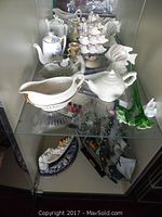 Various ceramic and nautical decor items arranged on glass shelving including gravy boat, teapots, ceramic ship and fish bowl.