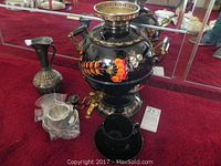 Photo showing Russian black tea pot with floral design, double handles, spout, black cup and saucer, metal pitcher and small metal vessel on red carpet