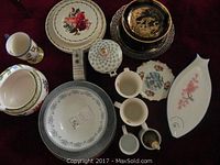 Mixed arrangement of floral dishes, serving platters, large mugs, decorative plates, and unique accent bowls on a red surface.