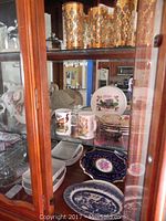 Photo showing display cabinet with various kitchenware items including gold-tone highball glasses, Dunoon coffee mugs, decorative plates and vintage Ronson lighter