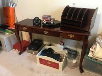 Photo of wooden desk with single drawer showing missing handle, letter organizer, clocks and assorted boxes underneath desk.