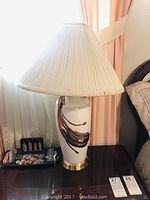 Two matching ceramic lamps with white pleated lampshades, gold swirl design on lamp body, and gold trim on base, placed on a dark wooden surface near window and curtains.
