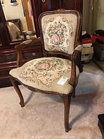 Full view of wooden arm chair showing floral and dancing couple tapestry upholstery on seat and backrest with carved wooden frame.