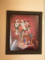 Framed still life artwork depicting a glass vase with pink and white flowers alongside vanity items on a table, dark ornate frame.