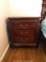 Front view of one nightstand showing three drawers with carved fronts and brass handles, dark wood finish, standing on carved feet.