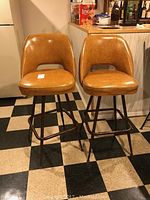 Front view of two mid century swivel bar stools with tan vinyl upholstery, metal legs, and footrests, on black and white checkered floor.
