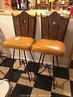Front view of two vintage bar stools with wood backs, tan upholstered swivel seats, and black metal frames, standing side by side on a checkered floor.