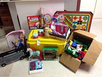 Wide photo showing the assorted toys including dolls, building blocks in box, a plastic horse, boxed board games, yellow storage tote and various small toys.