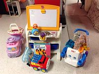 Photo of various children's toys including a purple and orange easel, pink push walker, potty seat, pegboard game, and toy firetruck.