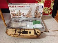 Photo of partially assembled wooden ship with hull and deck, displayed next to the original box showing completed model image.