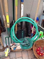 Photo showing garden tools including shovels, rakes, watering hose with nozzle, and small hand tools arranged against a wall.
