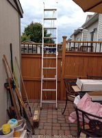 Full view of the extendable aluminum ladder standing upright on a patio with yard tools nearby.