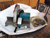 Front view of Makita Mitre Saw showing the base, motor housing, and blade with protective guard. Additional saw blades and accessories packaged beside it.