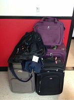 Luggage lot showing six different bags including purple, black, and gray suitcases and travel bags stacked together