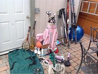 Wide shot of all sports equipment outside including skis, poles, golf bag, clubs, shoes, exercise items, badminton racket, and dartboard.