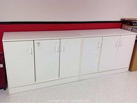 Two white cabinets placed side by side against a red and black wall. Both cabinets have two doors with white handles each, showing a clean front view.