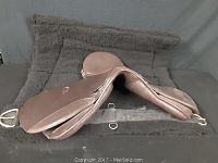 English saddle placed on a dark gray saddle pad, showing side profile with visible stitching, stirrup attachments, and padded seat.