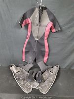 Photo showing the short sleeve wetsuit hung up with the zipper closed and a pair of black rubber open heel flippers in front on a black background.