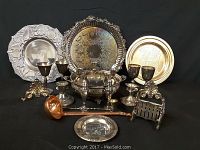 Full view of all silver-plated serving trays, goblets, covered serving dish, ladle, nutcracker, small dishes, and salt & pepper shakers displayed on black fabric background.