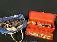 Photo showing a red metal toolbox with an open lid revealing various hand tools, a smaller red tray insert with additional tools in front, and a blue and white toolbag filled with assorted tools.