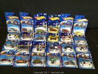 Top view of 19 Hot Wheels cars in original blister card packaging arranged in rows on black background.