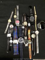 Photo showing full assortment of 16 watches laid out on black fabric surface, showcasing variety of styles, bands and colors.